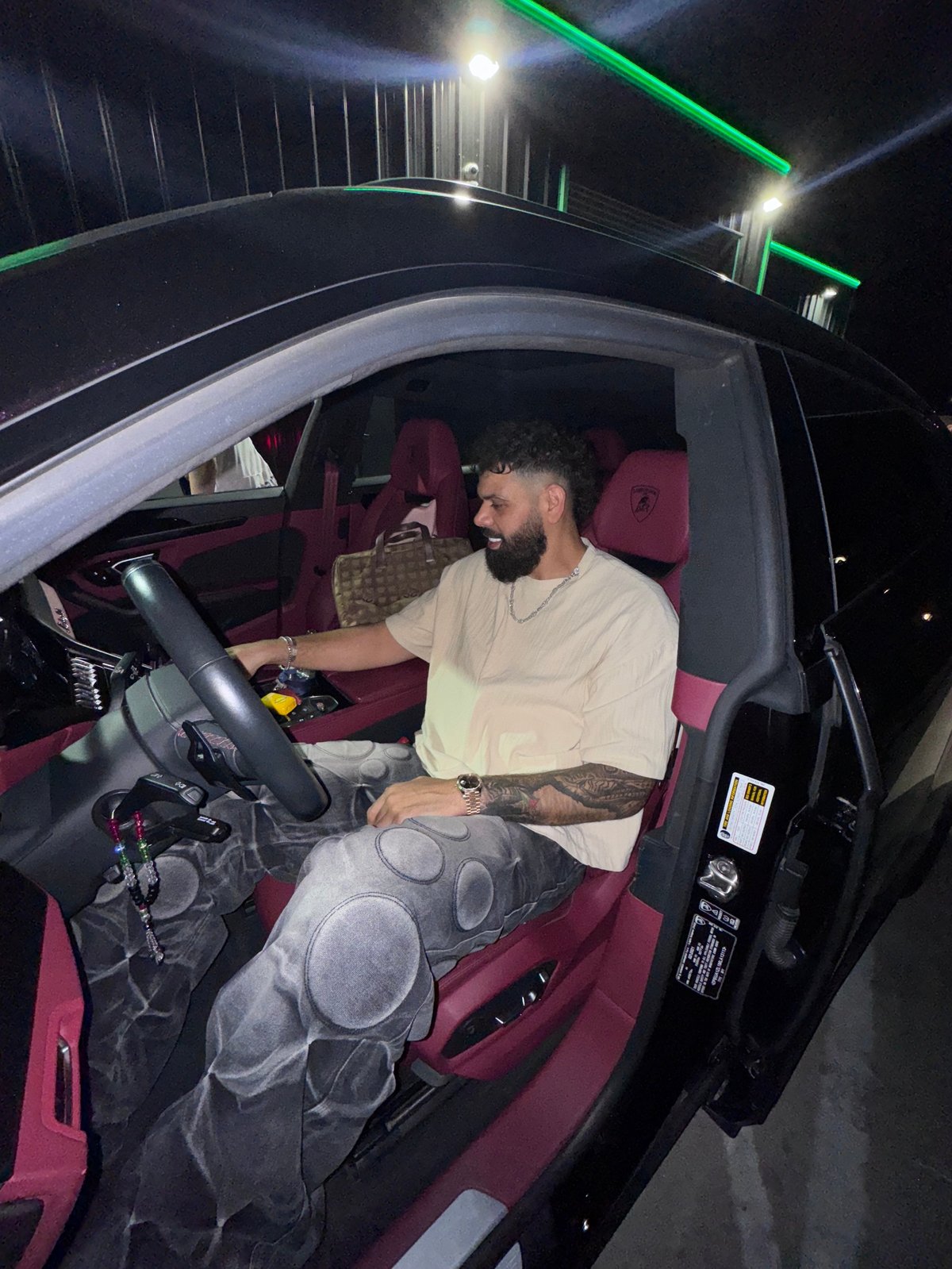 Bearded man with tattoo sitting in drivers seat of car with maroon interior at night, lit by green neon lights